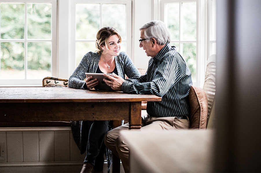 Senior man speaking with his adult daughter at his kitchen table while looking at a tablet.